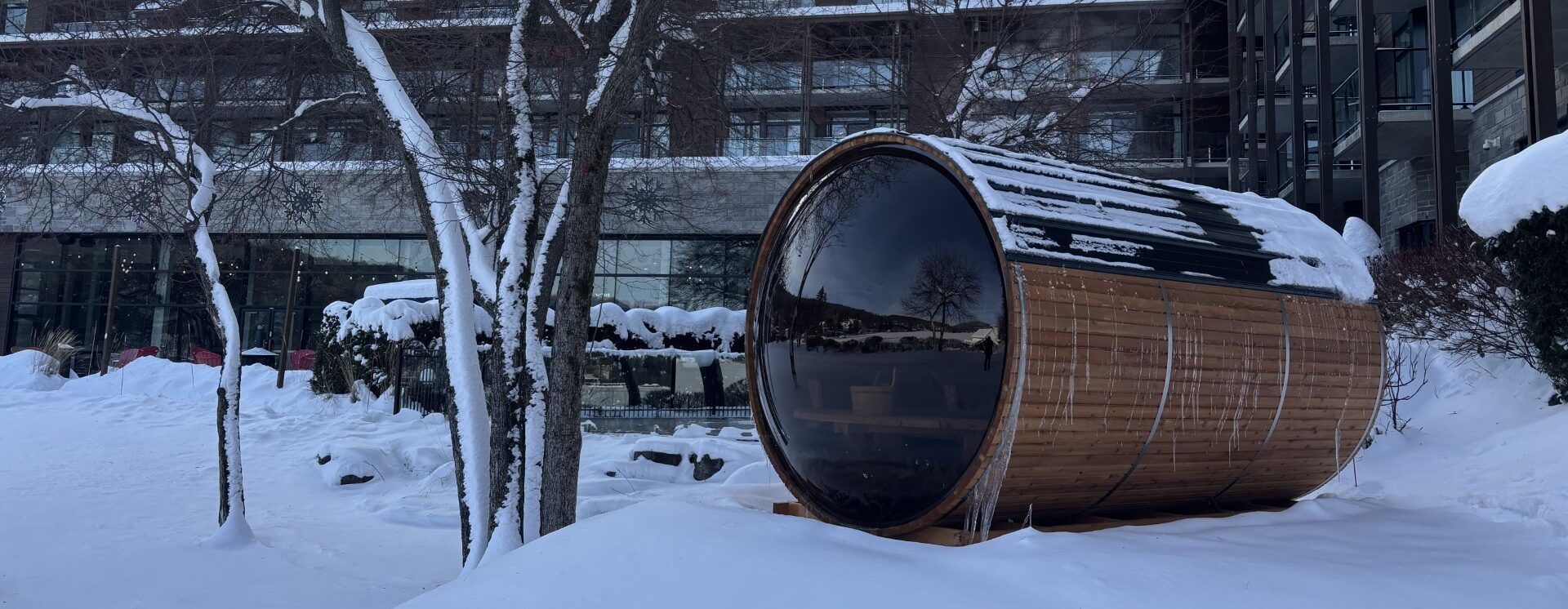 Sauna baril extérieur en cèdre avec façade vitrée installé dans un décor hivernal devant un complexe hôtelier