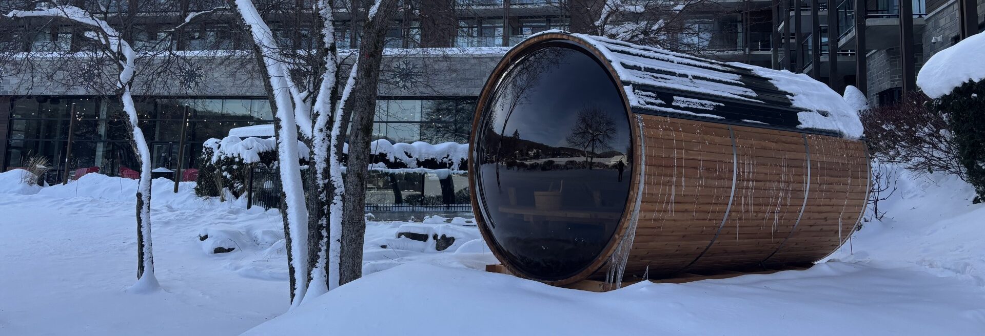 Outdoor cedar barrel sauna with panoramic glass front installed in a snowy resort setting during winter
