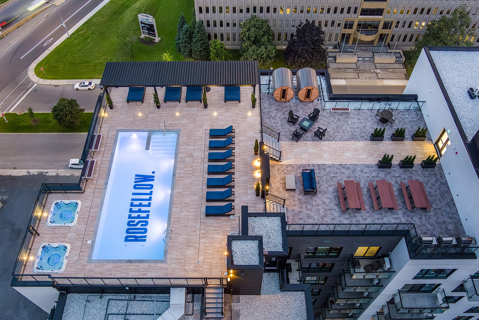 Aerial rooftop pool area with blue lounge chairs, two hot tubs, and a seating zone overlooking a city street.